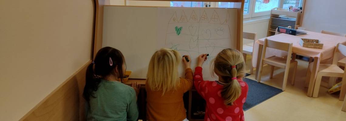 Drei Mädchen sitzen mit dem Rücken zum Betrachter. Sie sitzen vor einem Holzgestell mit leeren Blättern und malen auf einem Blatt gerade ein Bild. Im Hintergrund ist der Kindergartenraum mit weiteren Möbeln zu sehen.