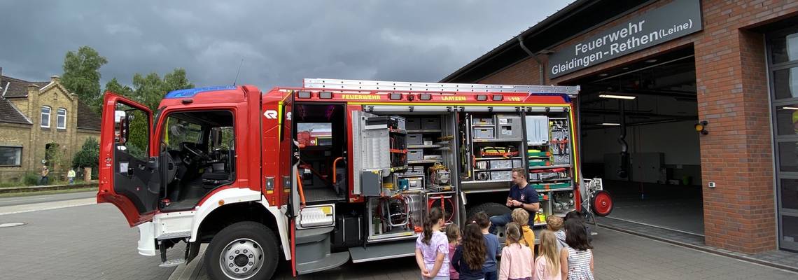 Das Bild zeigt eine Gruppe von Kindern, die vor einem geöffneten Feuerwehrauto stehen und einem Feuerwehrmann zuhören. Sie befinden sich vor der Feuerwache Gleidingen-Rethen (Leine). Das Auto ist mit vielen Geräten ausgestattet. Der Himmel ist bewölkt.