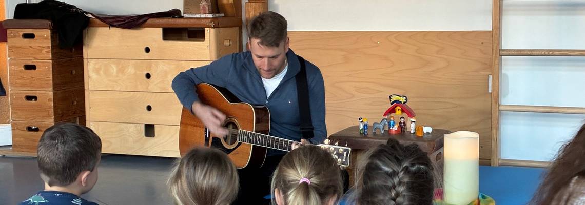 Im Vordergrund sind Kinder zu sehen. Sie sitzen mit dem Rücken zur Kamera auf Blauen Matten auf dem Boden. Im Mittelpunkt des Bildes ist ein Mann über seine Gitarre gebeugt. Neben dem Mann steht auf einem kleinen Hocker eine Krippenszene, daneben steht eine große weiße Kerze und ein buntes Tuch.