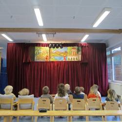 Kindergruppe sitzt auf kleinen Stühlen mit dem Blick zum Theaterstück. Eingerahmt von einem roten Vorhang ist eine Szene aus dem Stück "Backe, backe Kuchen" zu sehen..