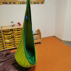 Ein kleiner Raum. auf dem Boden liegt ein orangener Teppich. im Mittelpunkt ist eine Schaukel aus grünem Stoff zu sehen.