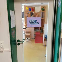 Tür des Gruppenraums. In einem grünen Türrahmen ist eine weiße Tür mit großer Glasfläche eingelassen. Auf der Glasscheibe ist ein Bild mit der Aufschrift "Rein ins Bunte Vergnügen" zu sehen.