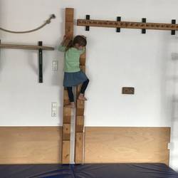 Ein Kind klettert an senkrecht an der Wand befestigten Stufen aus Holz. Diese beginnen an einer, an der Wand befestigten, Kletterstrecke aus Holz- und Metalelementen und enden auf einer Weichbodenmatte.