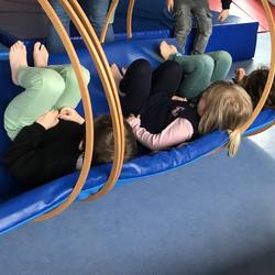 Drei Kinder liegen in einer Wippe. Diese besteht aus einer dünnen blauen Matte die in mehrere Holzreifen gelegt ist.