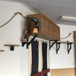 Eine Kletterstrecke an der Wand ist zu sehen. Diese besteht aus an der Wand befestigten Seilen zum Festhalten, verschieden Trittstufen auf Holz und Metal und einem Tunnel aus Holz zum durchklettern.  Ein Kind ist von hinten zu sehen, das in die Öffnung des Tunnels klettert.