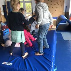Mehrere Kinder sind von hinten zu sehen, die auf einer großen, blauen Weichbodenmatte stehen.