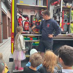 Ein Feuerwehrmann zeigt einer Kindergruppe die Ausrüstung im Feuerwehrauto. Ein Mädchen steht direkt daneben und schaut interessiert zu. Zu sehen sind Feuerlöscher, Werkzeuge und Geräte.