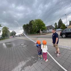 Zwei Kinder mit orangenen Helmen spritzen mit einem Feuerwehrschlauch Wasser auf einen Platz. Ein Feuerwehrmann steht daneben und beobachtet sie. Der Himmel ist bewölkt.