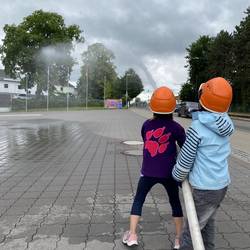 Zwei Kinder mit orangenen Helmen spritzen mit einem Feuerwehrschlauch Wasser auf einen Platz. Der Himmel ist bewölkt.