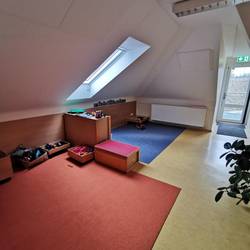 Ein Raum unter der Dachschräge ist zu sehen. Auf dem Boden liegen zwei Teppiche in Rot und Blau. Auf den Teppichen sind Kisten mit verschiedenen Materialien.