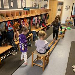 Im Mittelpunkt des Bildes sind drei Kinder zu sehen. Das Erste balanciert über zwei dünne Holzbalken, die zwei Holzhocker miteinander verbinden. Das Zweite sitzt auf dem Holzhocker und wartet. Daneben steht das dritte Kind.