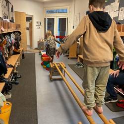 Ein Kind im Vordergrund balanciert über zwei dünne Holzbalken. Im Hintergrund hüpft ein anderes Kind über bunte halbrunde Steine.