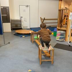 Im Mittelpunkt des Bildes ist ein Kletterelement zu sehen. es besteht aus zwei Holz Hockern mit Stoff Sitzfläche und einer Holzplanke dazwischen. Ein Kind stellt sich freihand auf dem vorderen Hocker auf.