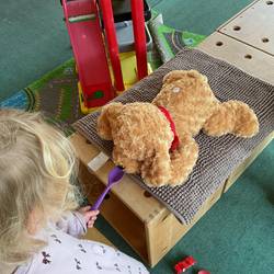 Ein Kind kniet auf dem Boden und hält einen leeren Löffel vor die Schnauze eine Stofftier Hundes. Der Stofftier Hund liegt auf dem Bauch..