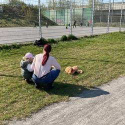 Eine Fachkraft und ein Kind sind von hinten fotografiert. Sie knien auf einer Rasenfläche. Vor ihnen liegt ein Stofftier Hund. Im Hintergrund sind spielende Kinder auf einem asphaltierten Bolzplatz zu sehen.