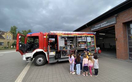 Das Bild zeigt eine Gruppe von Kindern, die vor einem geöffneten Feuerwehrauto stehen und einem Feuerwehrmann zuhören. Sie befinden sich vor der Feuerwache Gleidingen-Rethen (Leine). Das Auto ist mit vielen Geräten ausgestattet. Der Himmel ist bewölkt.