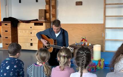 Im Vordergrund sind Kinder zu sehen. Sie sitzen mit dem Rücken zur Kamera auf Blauen Matten auf dem Boden. Im Mittelpunkt des Bildes ist ein Mann über seine Gitarre gebeugt. Neben dem Mann steht auf einem kleinen Hocker eine Krippenszene, daneben steht eine große weiße Kerze und ein buntes Tuch.